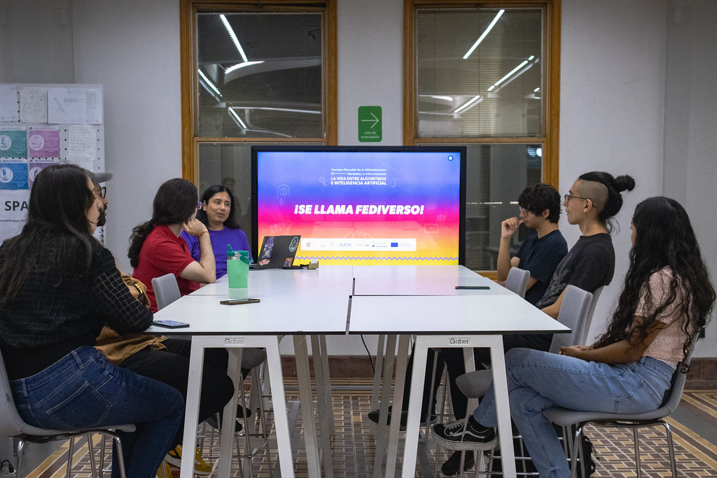 Mesa alta con siete personas sentadas alrededor en el Área de Producción Digital del LabNL, al fondo y al centro hay una pantalla con el nombre de la sesión: ¡Se Llama Fediverso!