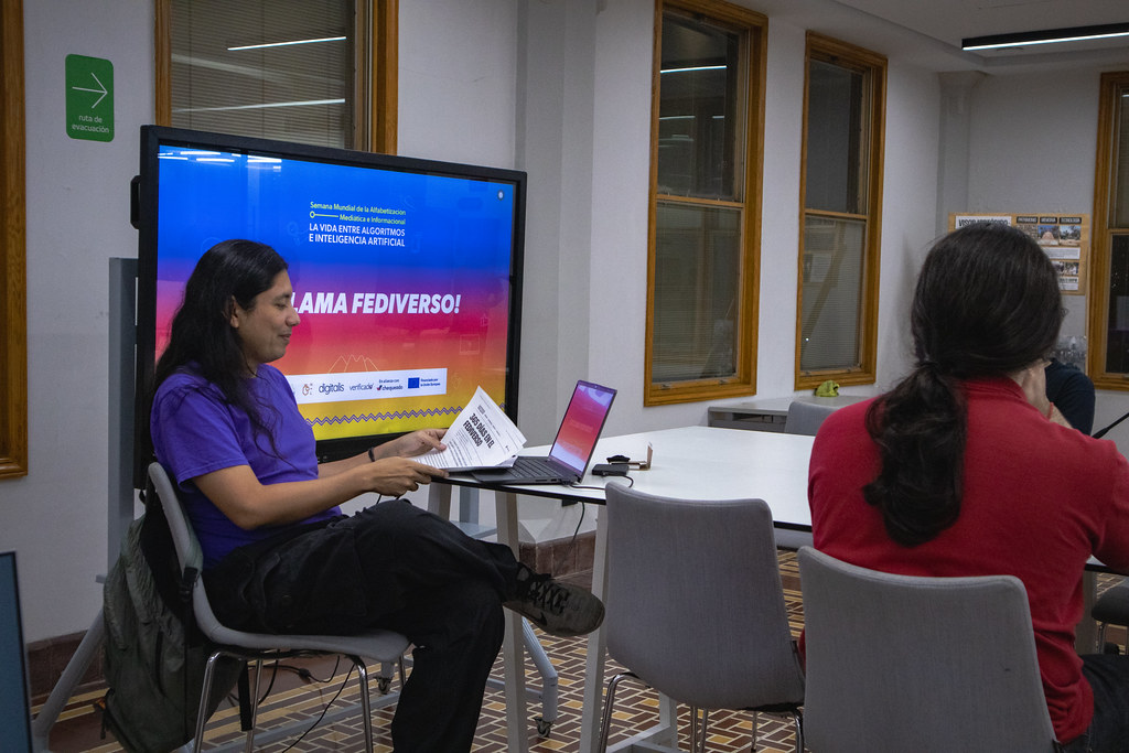 Plano medio de una mesa alta con tres personas sentadas alrededor en el Área de Producción Digital del LabNL, al fondo y a la izquierda hay una pantalla con el nombre de la sesión: ¡Se Llama Fediverso!