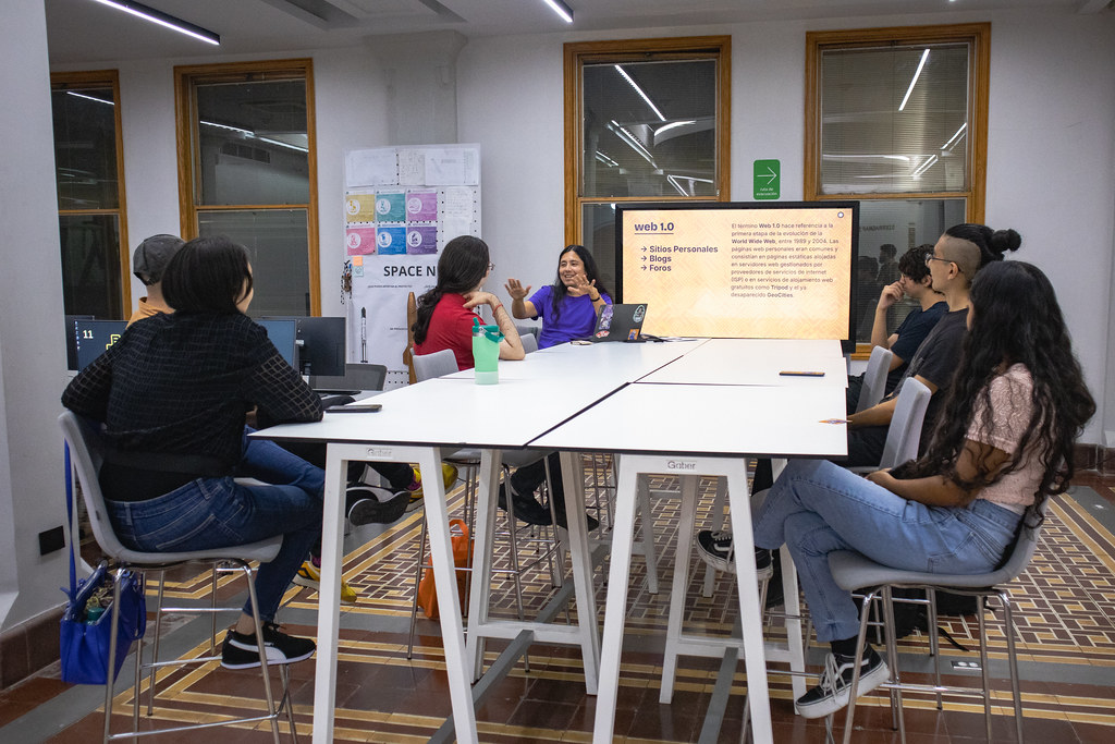 Mesa alta con siete personas sentadas alrededor en el Área de Producción Digital del LabNL, al fondo y al centro hay una pantalla con el nombre de la sesión: ¡Se Llama Fediverso!
